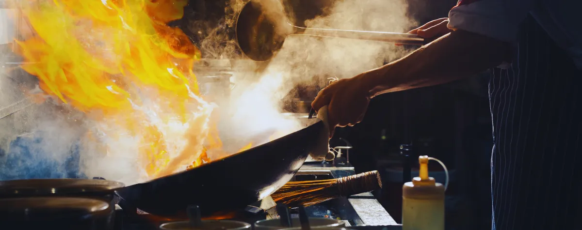 A chef cooking with a frying pan over an open flame within a restaurant kitchen