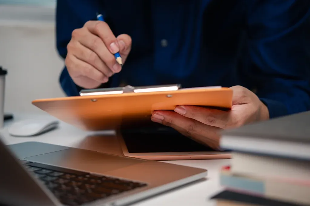 Person writing on a clipboard at a desk with a laptop and office documents nearby.